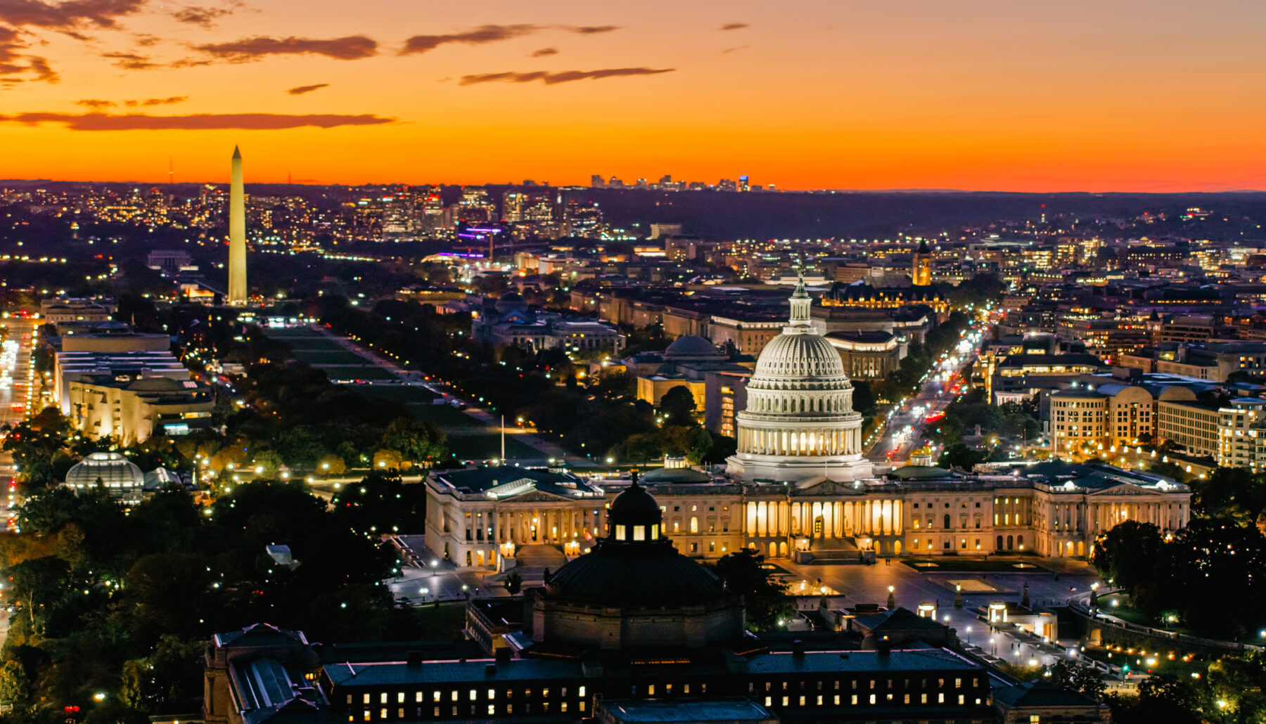 Aerial view of Washington, DC at dusk showing the illuminated US Capitol building in the centre, with the Washington Monument visible in the background to the left. The city lights glow against an orange and purple sunset sky.