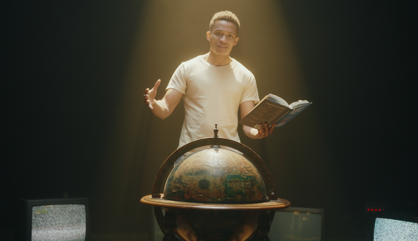 Aric standing behind a globe holding a book, still from a recent video